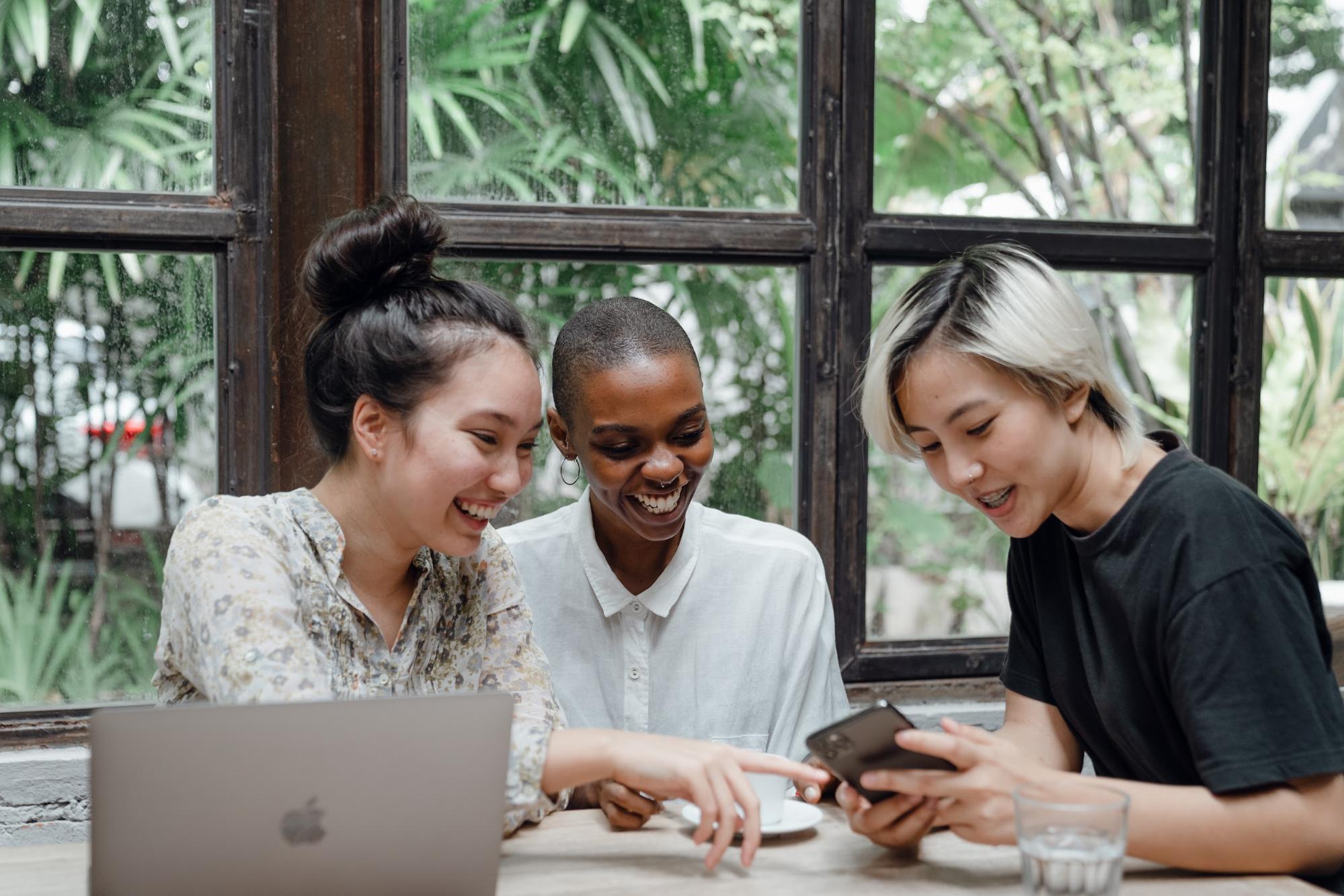 Three young women sitting at a coffee shop looking delighted by a phone.